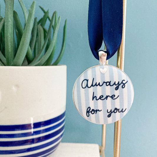 A blue and white striped fabric keepsake with the words Always Here For You, on a navy blue ribbon