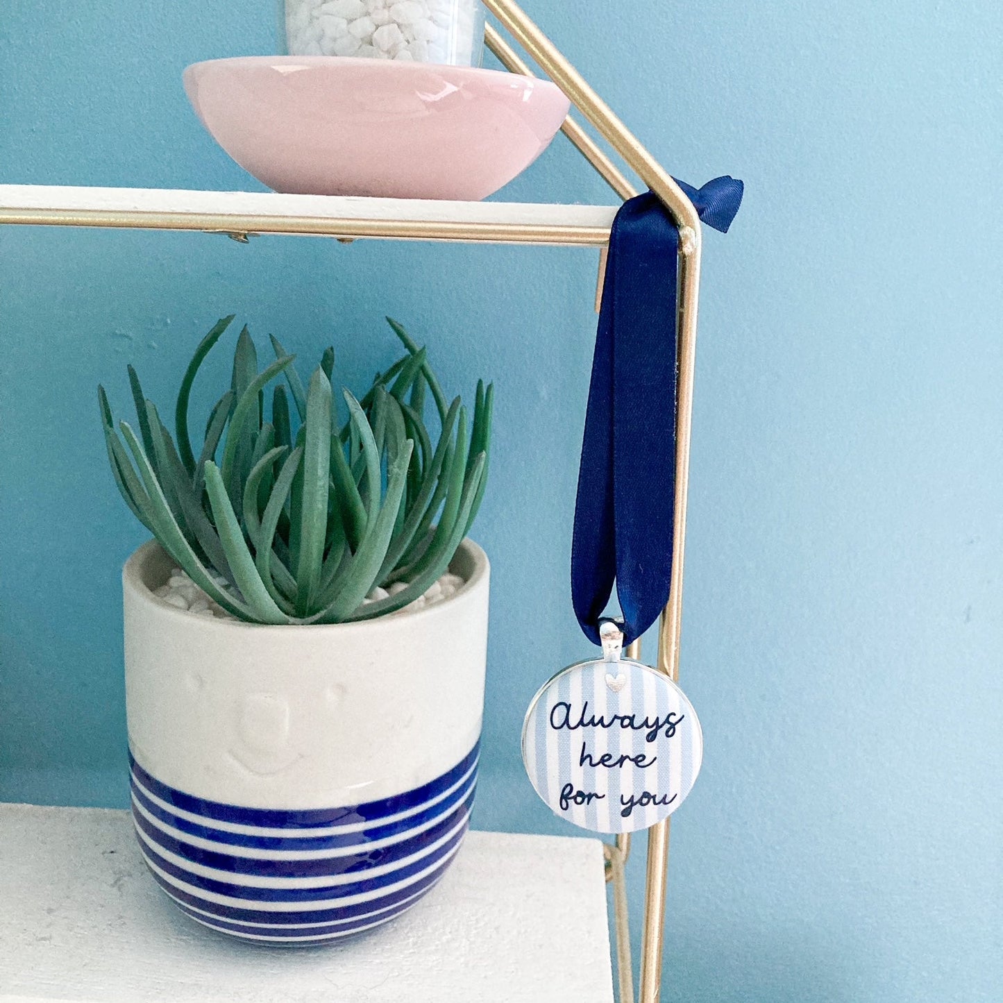 A blue and white striped fabric keepsake with the words Always Here For You, on a navy blue ribbon