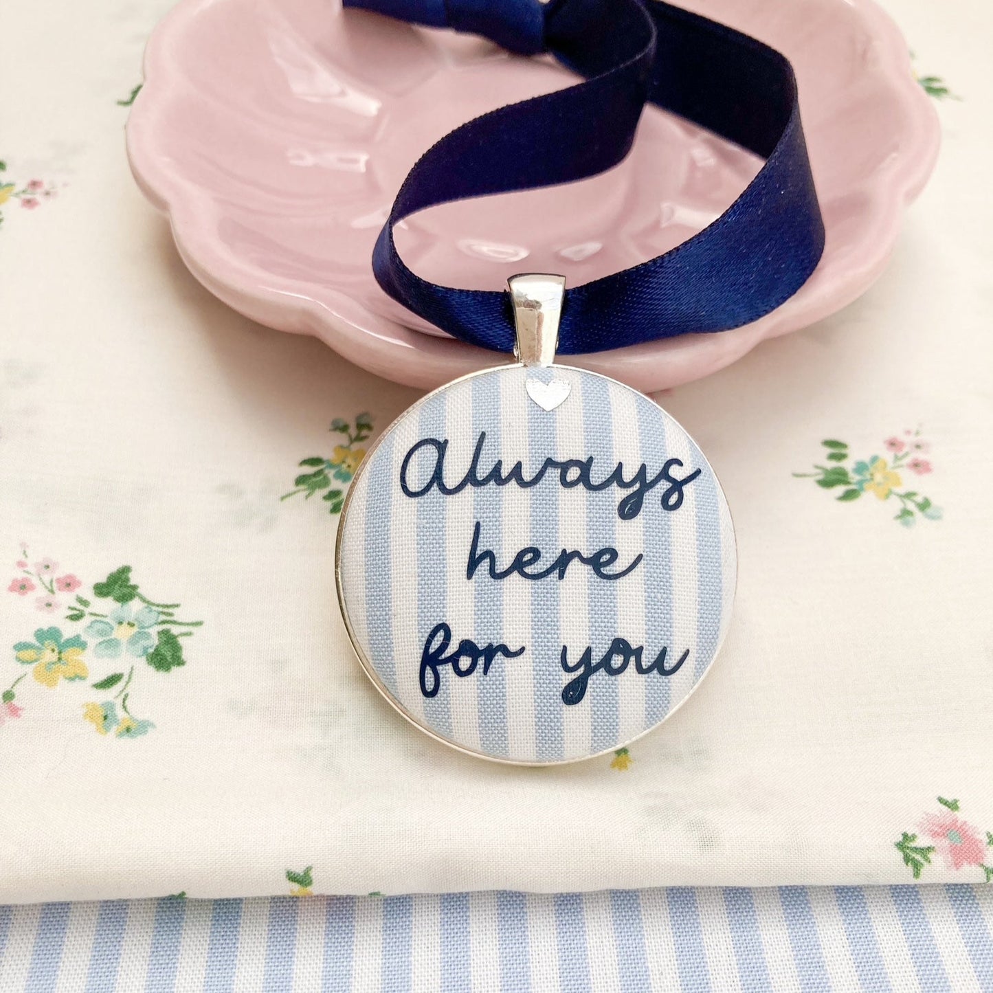 A blue and white striped fabric keepsake with the words Always Here For You, on a navy blue ribbon