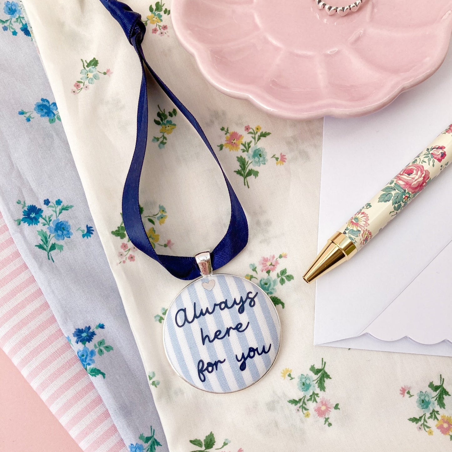 A blue and white striped fabric keepsake with the words Always Here For You, on a navy blue ribbon