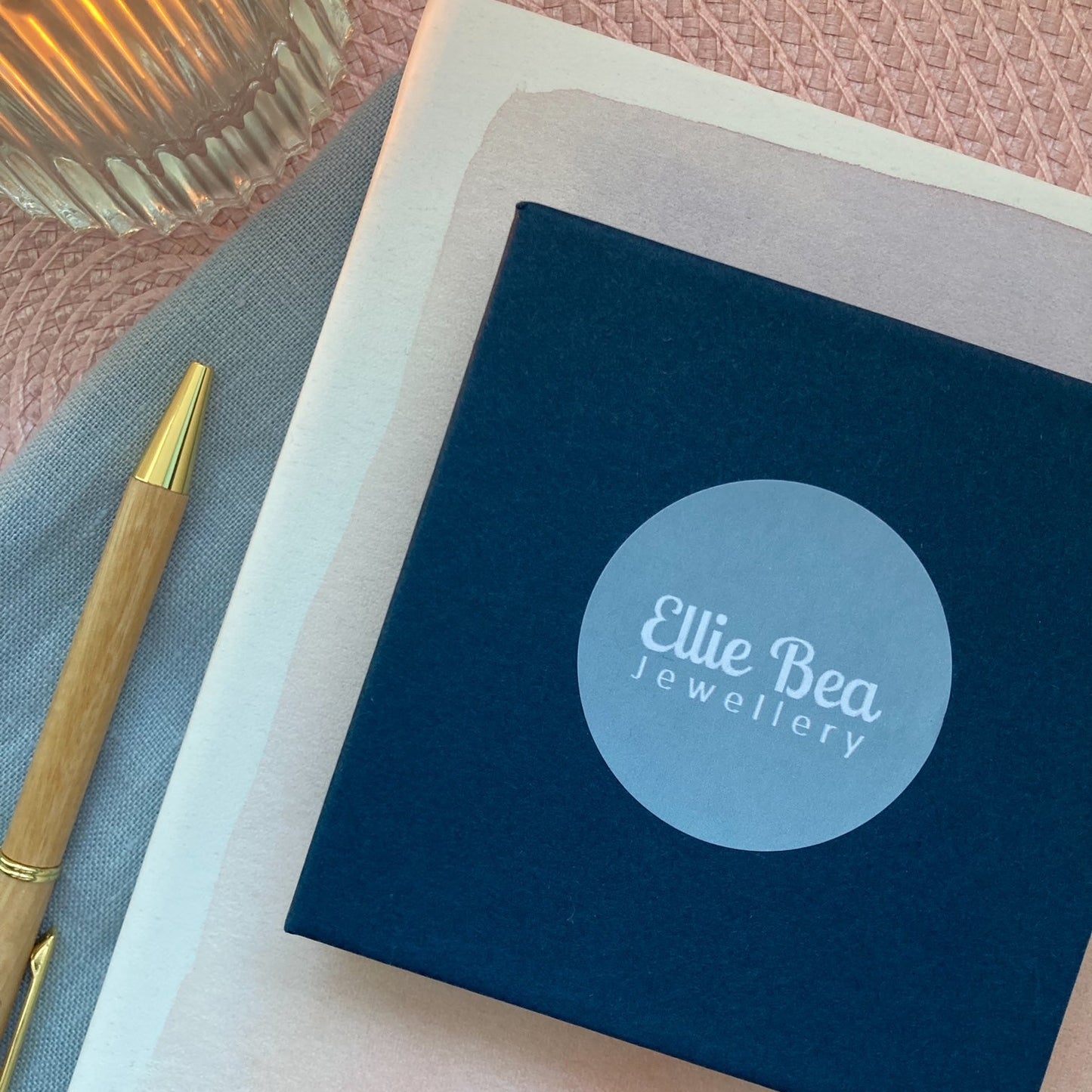 Blue box with 'Ellie Bea Jewellery' logo on a pink textured surface with a pen and candle.