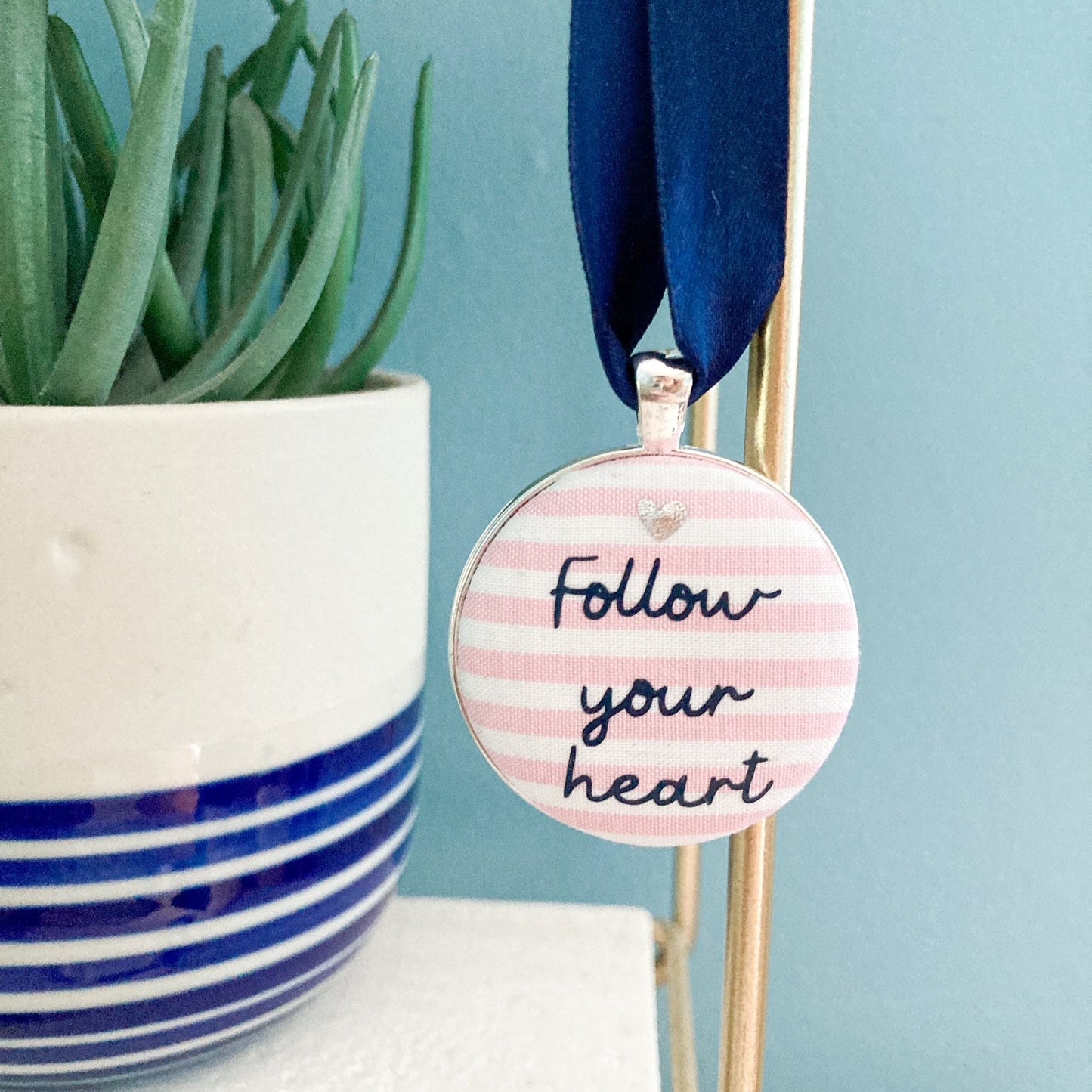 Keepsake with Follow Your Heart printed onto pink & white striped fabric, hanging on a navy blue ribbon