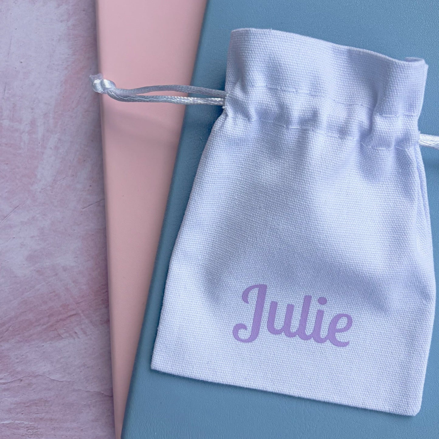 A small white drawstring pouch with the name 'Julie' printed in purple script, placed on top of a dusty blue notebook and a pink book on a textured pink surface.