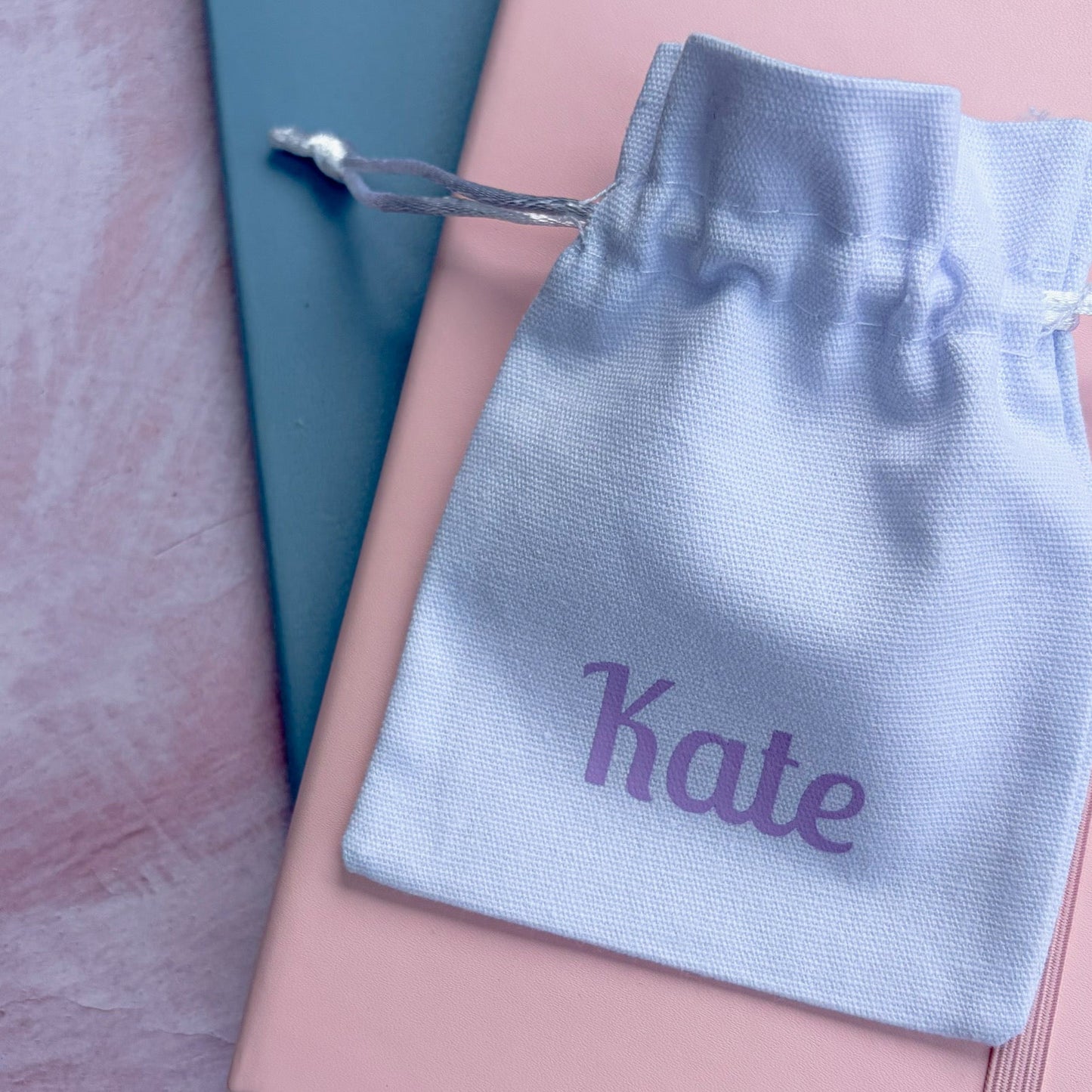 A light blue personalized drawstring pouch printed with the name 'Kate' in purple, resting on top of a pink notebook and a teal book on a textured pink surface.