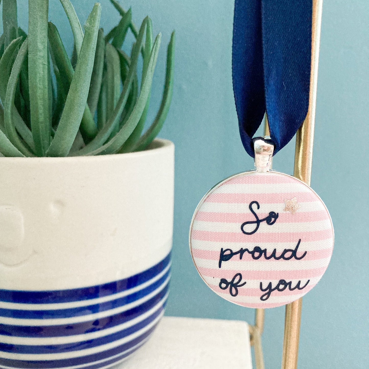 Keepsake with So Proud Of You printed onto pink and white striped fabric with a navy blue ribbon