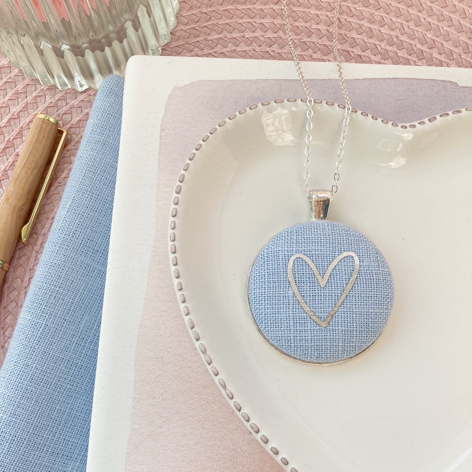 Heart-shaped jewelry dish with a necklace featuring a heart pendant on a pink and blue fabric background.