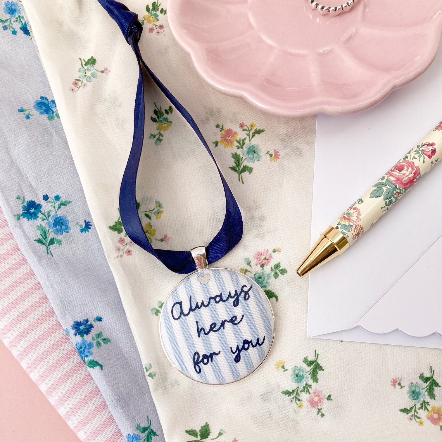 A blue and white striped fabric keepsake with the words Always Here For You, on a navy blue ribbon