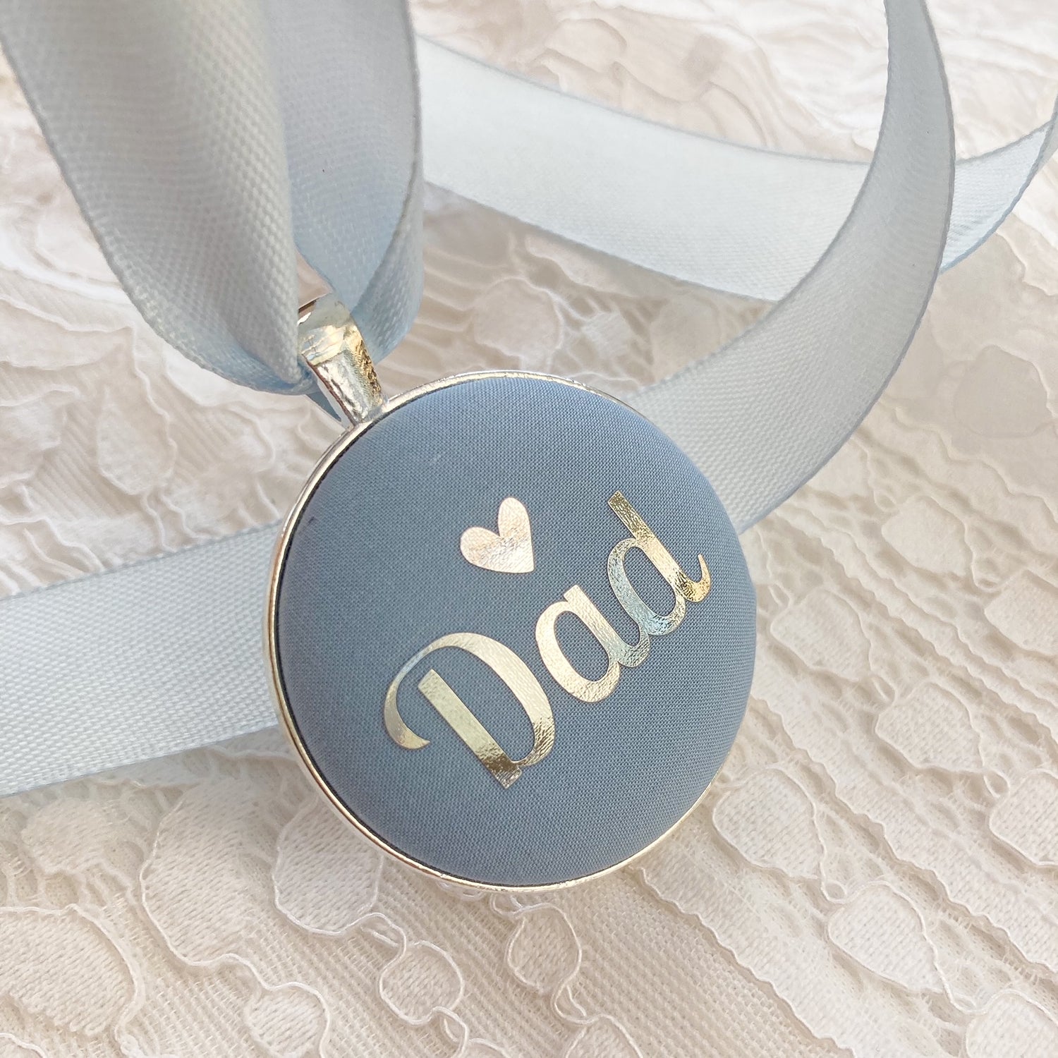 A round light blue wedding bouquet charm with metallic silver foil lettering that reads 'Dad' and a small heart, attached to a pale blue ribbon and photographed on a white lace fabric background.
