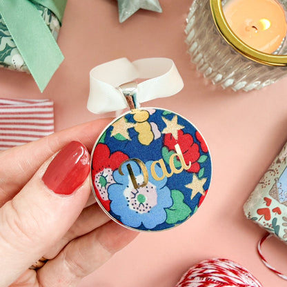 Personalised Christmas keepsake or memory keepsake with dad printed in gold foil, made with blue floral and star Liberty fabric, in a silver plated setting, thread on white satin ribbon, on a Christmas flatlay