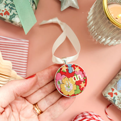 A Christmas personalised keepsake or memory keepsake, with gran printed in gold foil, on Liberty red floral fabric, thread on an ivory ribbon, and set on a Christmas flatlay