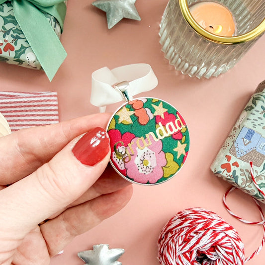 A Grandad Christmas keepsake or memory keepsake, made with green floral Liberty fabric, printed with Grandad in gold foil, set in silver plate, thread on a white satin ribbon, on a Christmas flatlay