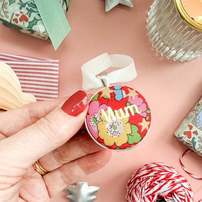 A Christmas Keepsake gift or memory keepsake, made with red floral Liberty fabric, printed with Mum in gold, thread on an ivory white ribbon and set on a Christmas flatlay