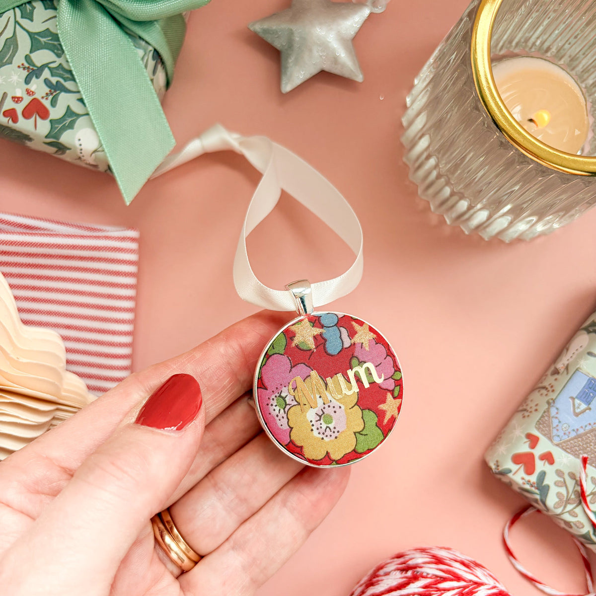 A Christmas Keepsake gift or memory keepsake, made with red floral Liberty fabric, printed with Mum in gold, thread on an ivory white ribbon and set on a Christmas flatlay