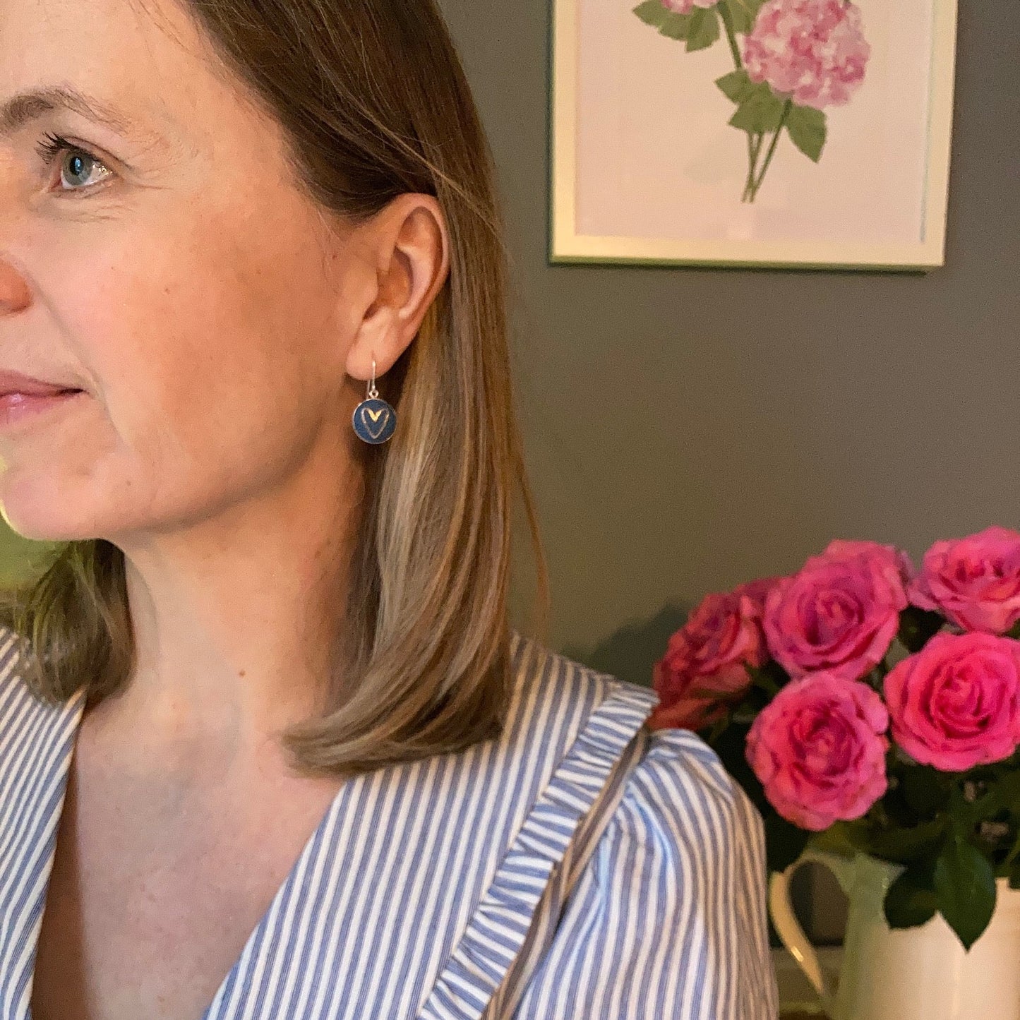 Woman wearing a blue and white striped shirt and blue heart earrings with a floral arrangement in the background