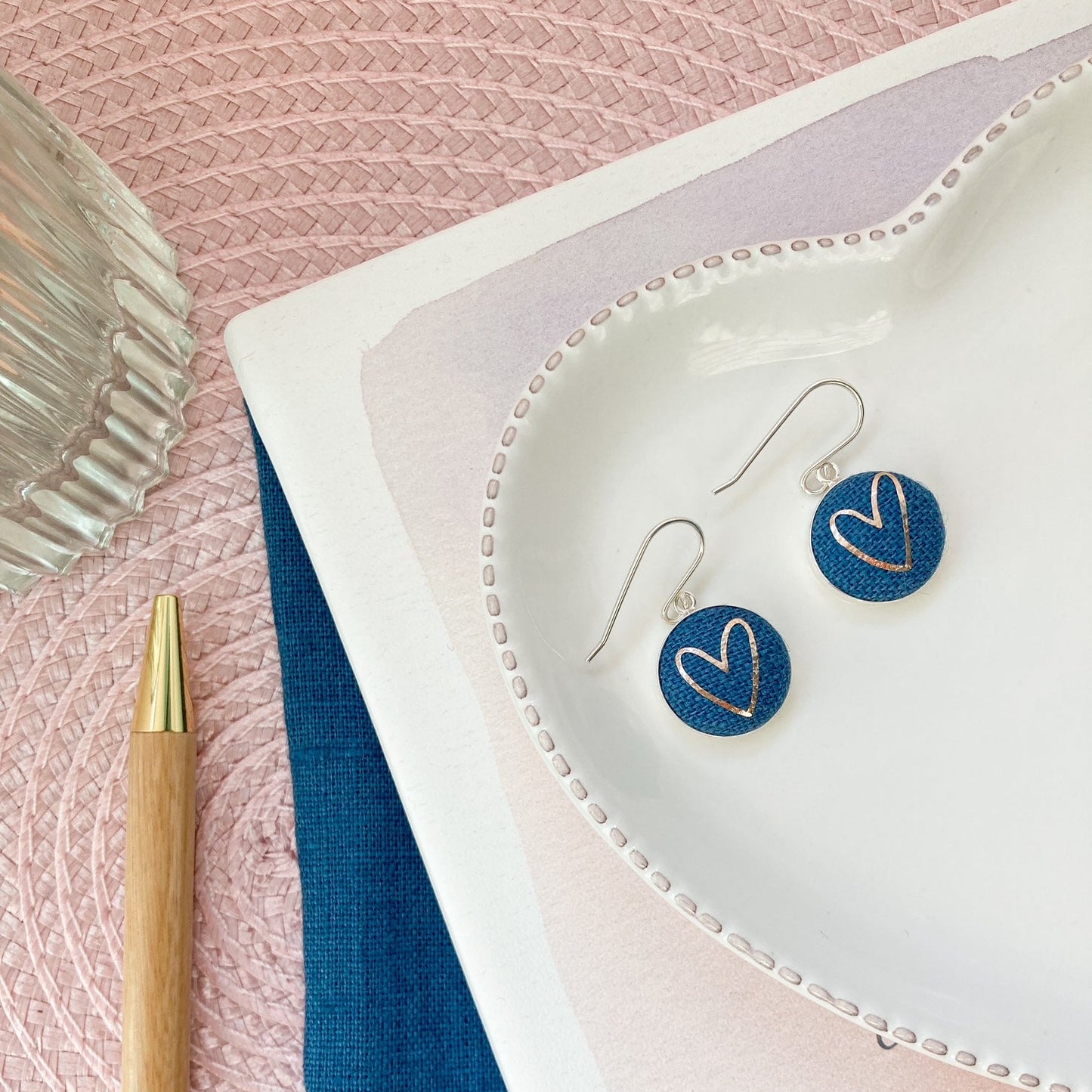 Heart-shaped earrings on a white dish with a pen and fabric on a pink surface