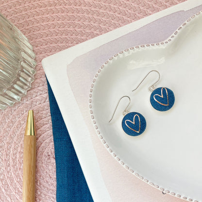 Heart-shaped earrings on a white dish with a pen and fabric on a pink surface