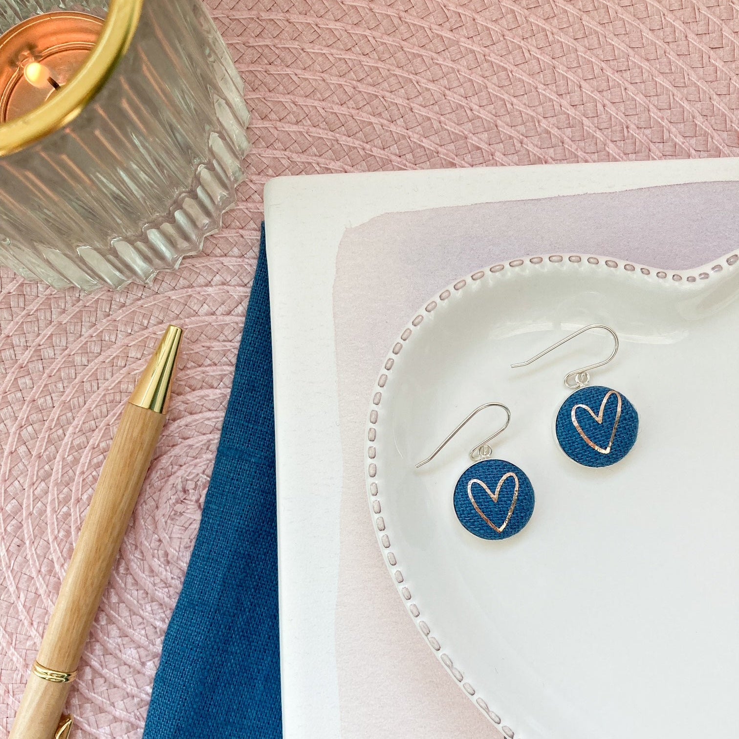 Heart-shaped earrings on a white dish with a pen and candle on a pink fabric background