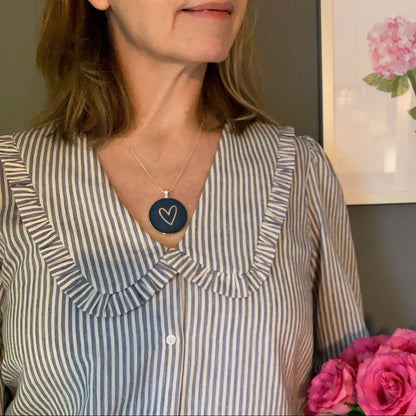 Woman wearing denim blue and gold heart necklace, holding pink roses with a framed picture of flowers on the wall.