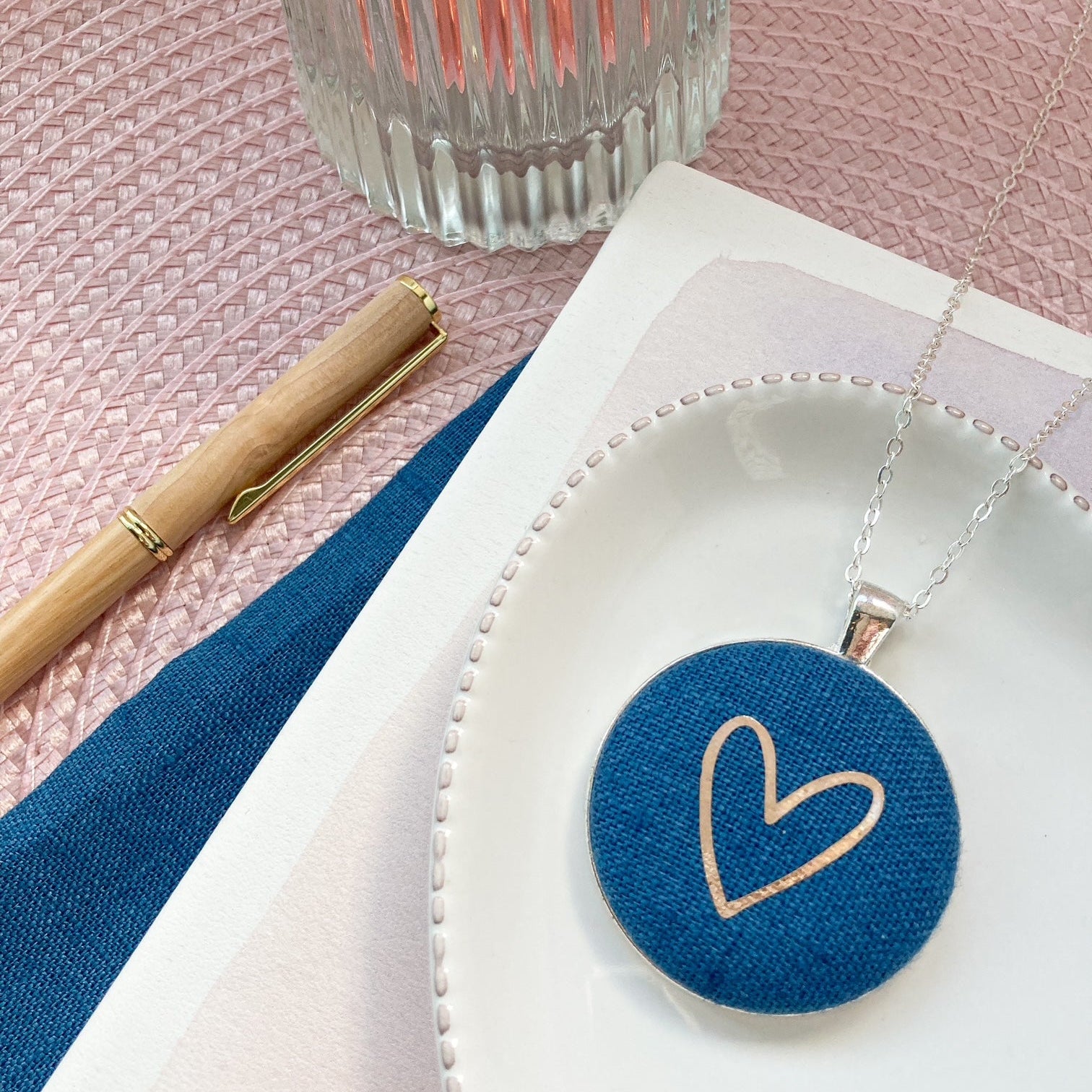 Blue and gold heart pendant on a white dish with a pink tablecloth and candle in the background