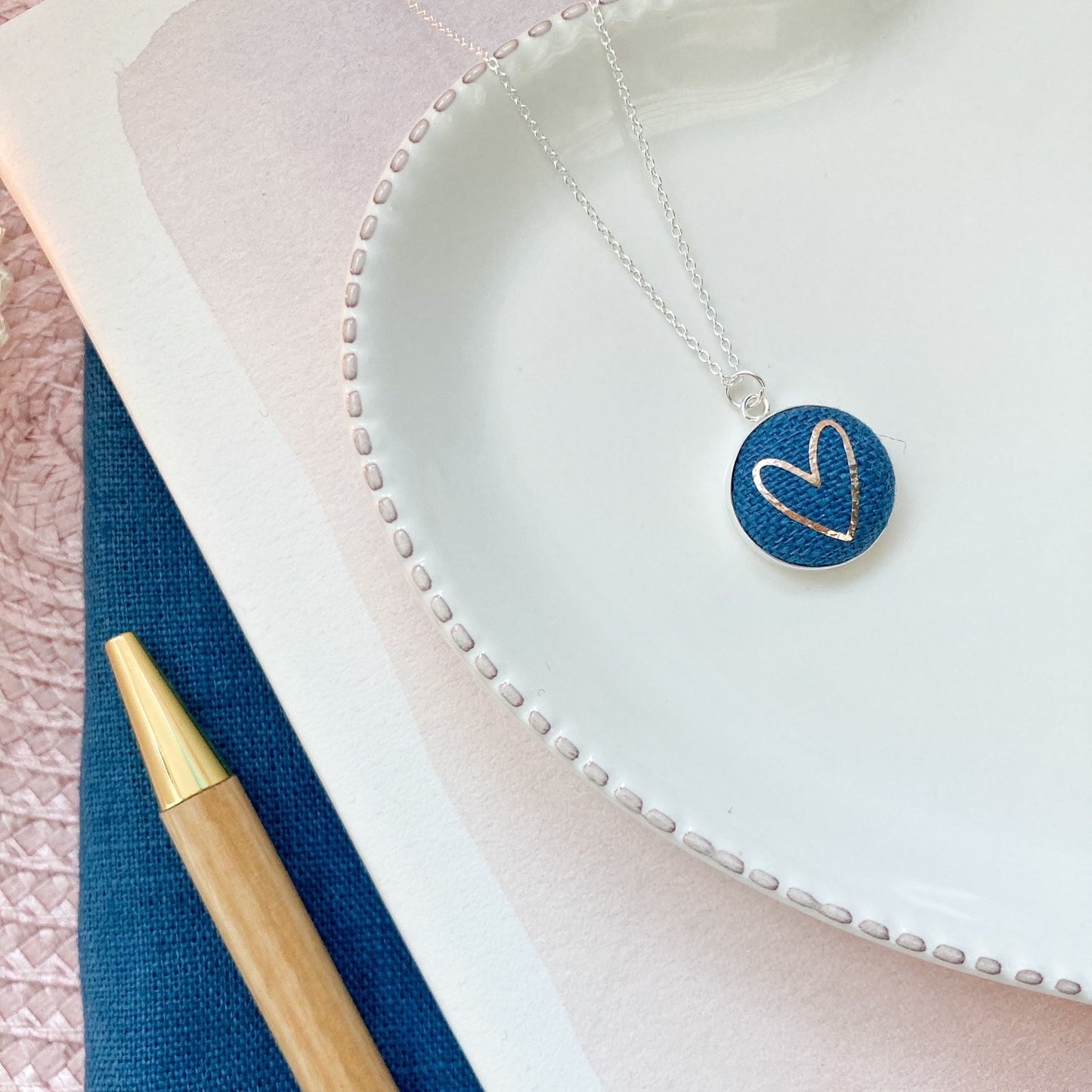 Necklace with a blue heart pendant on a white surface with a pen next to it.