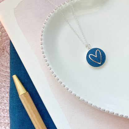 Necklace with a blue heart pendant on a white surface with a pen next to it.