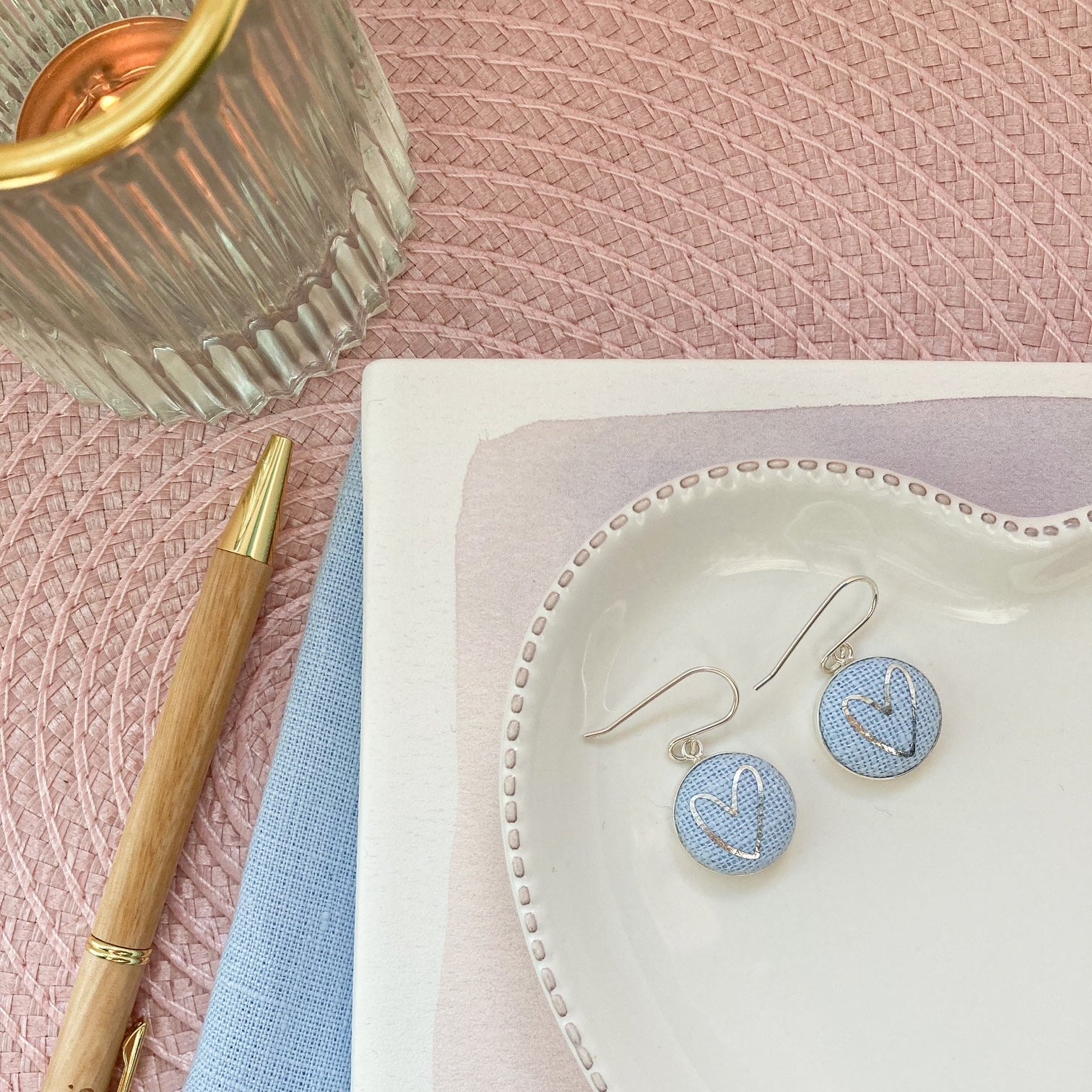 Earrings on a white dish with a pen and glass on a pink textured surface