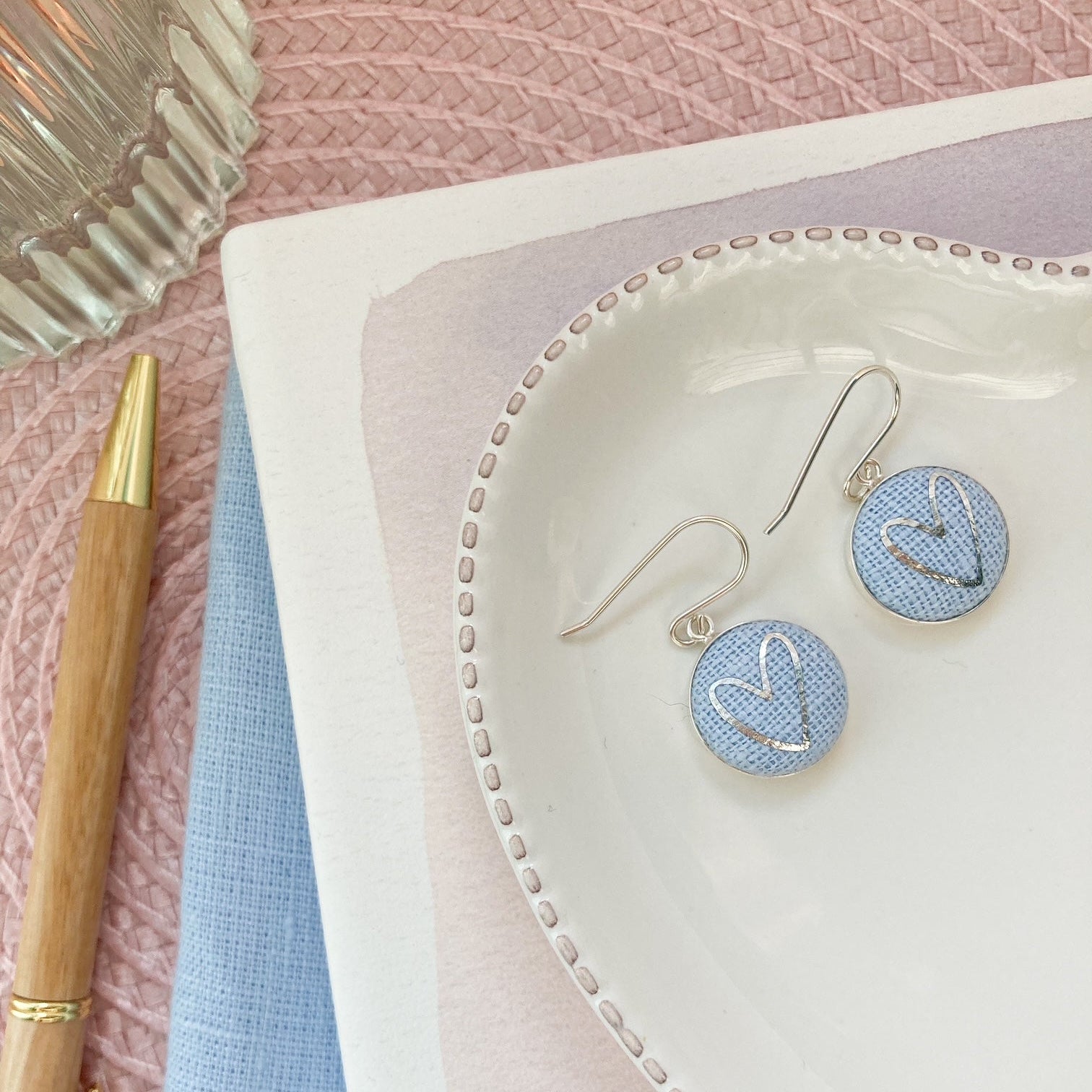 Earrings on a heart-shaped dish with a pen and fabric in the background