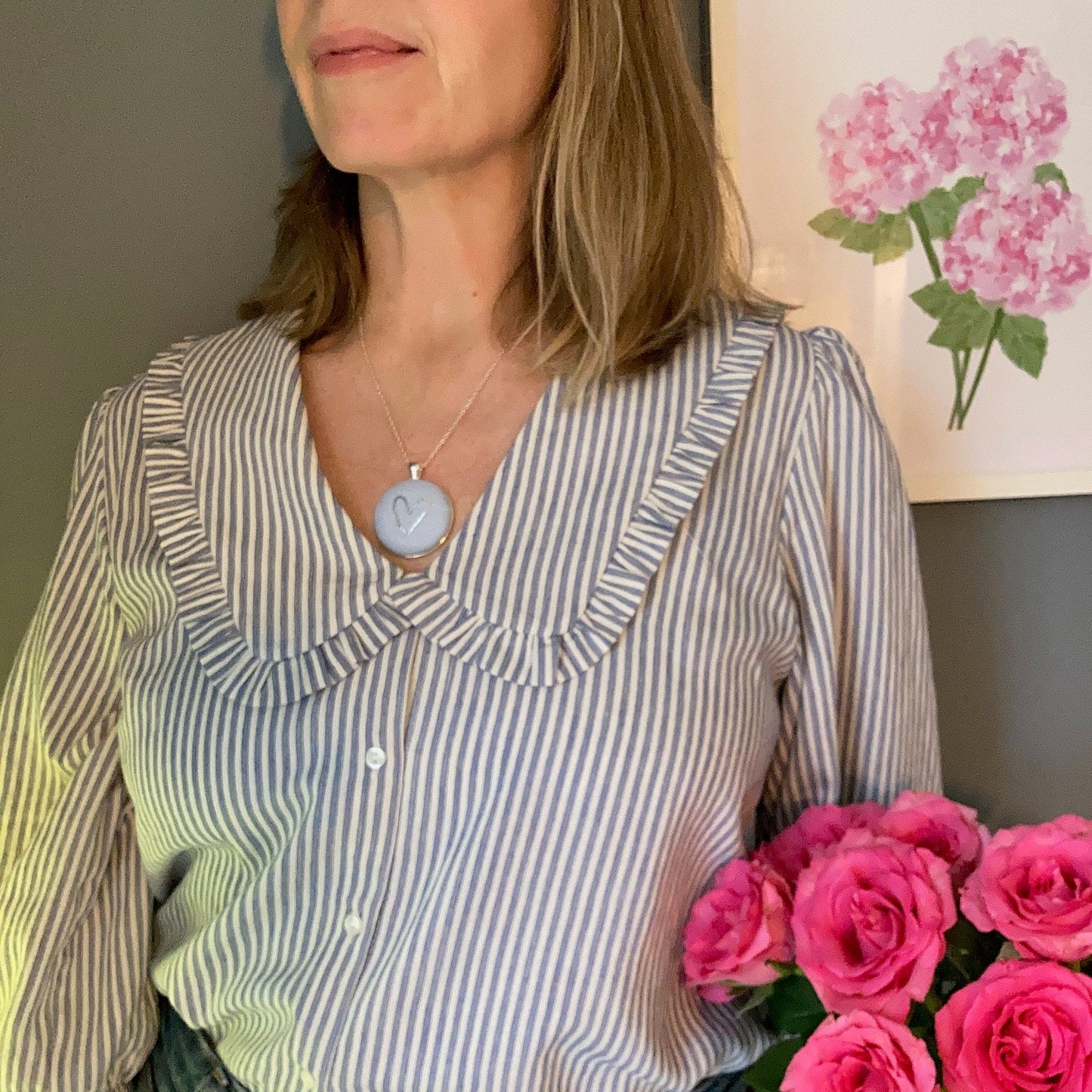 Person wearing a blue heart necklace and striped shirt with pink flowers and a painting in the background