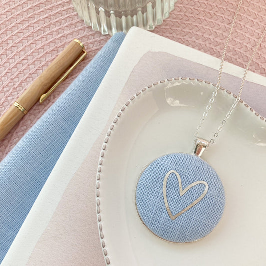 Necklace with a heart pendant on a pink surface with a pen and notebook.