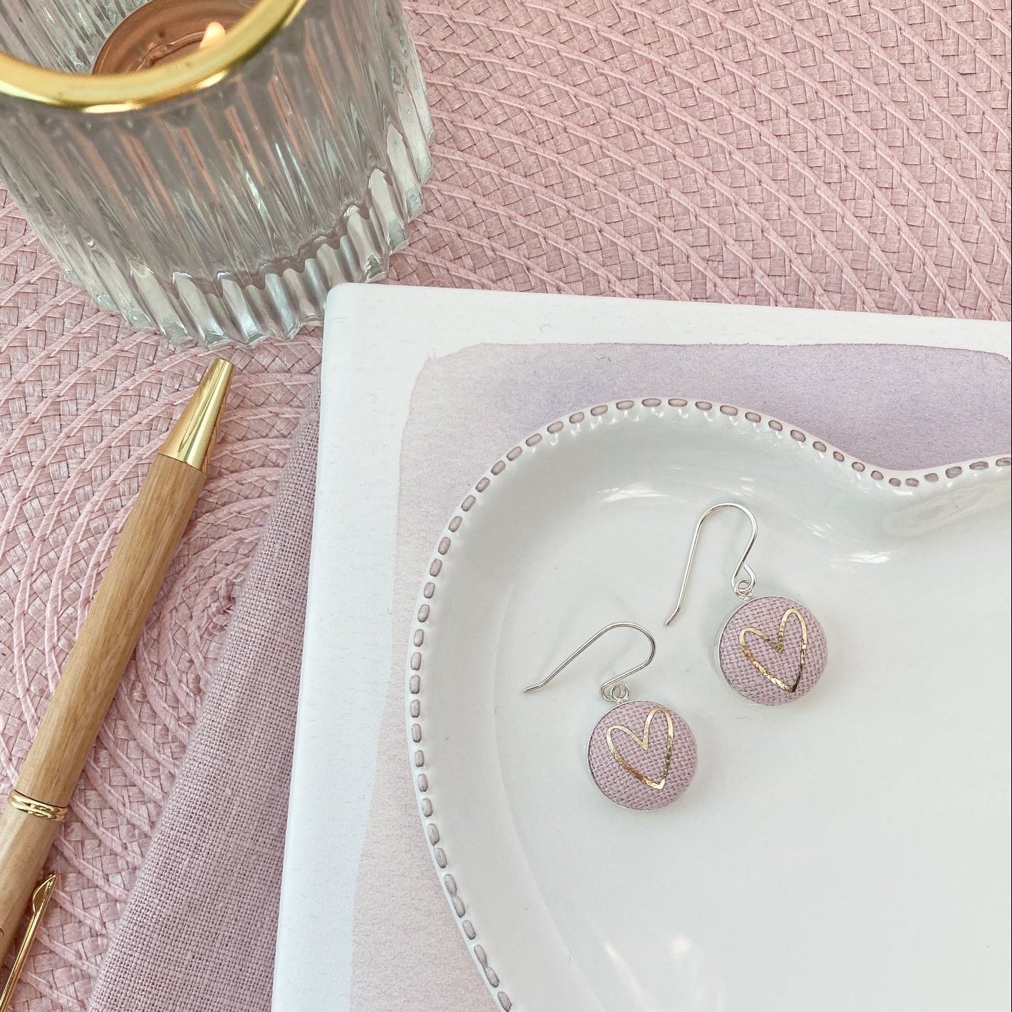 Heart-shaped jewelry dish with pink earrings on a pink textured surface with a pen and glass.