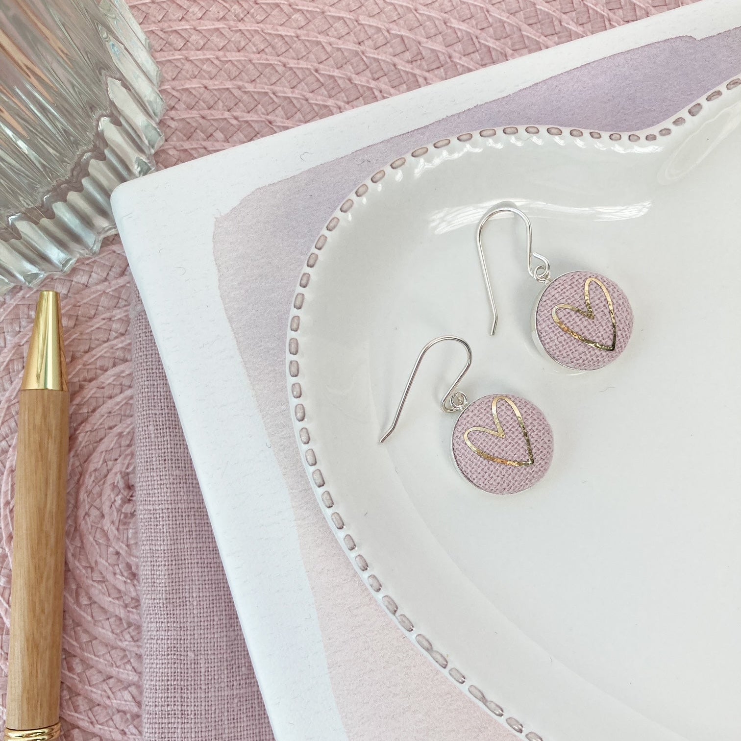 Pink and silver earrings with heart designs on a white dish with a pink textured background