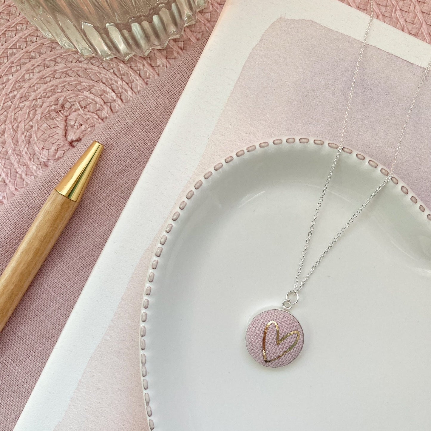 Necklace with a pink pendant on a white dish, next to a pen and textured pink surface.