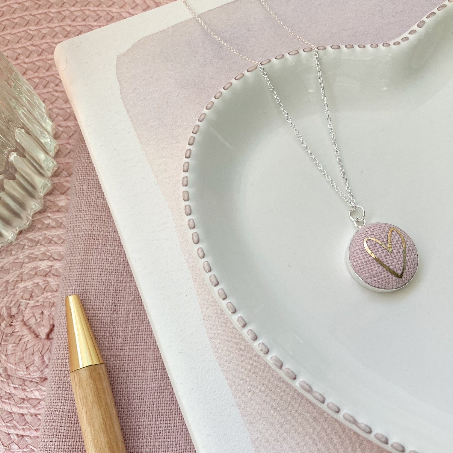 Necklace with a heart-shaped pendant on a pink textured surface with a pen.
