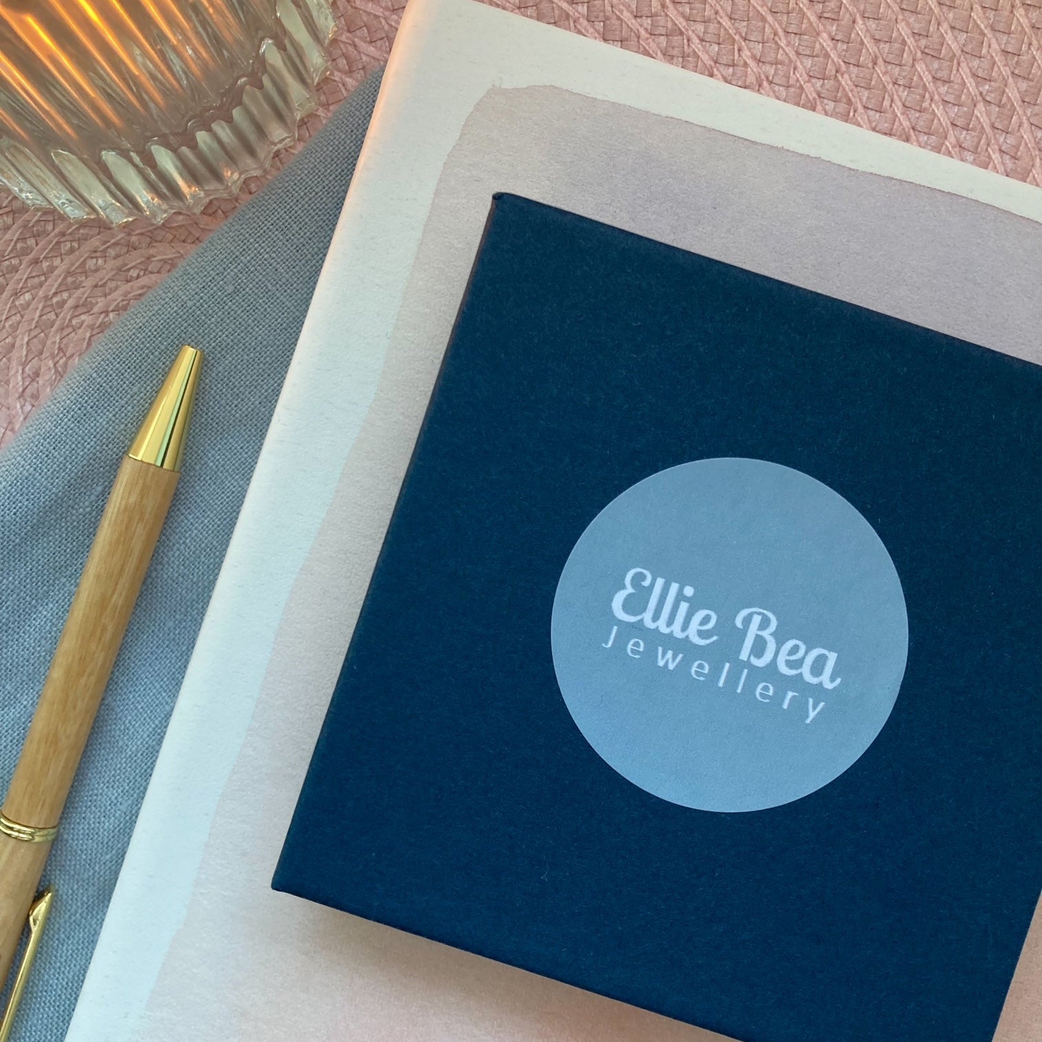 Blue box with 'Ellie Bea Jewellery' logo on a pink textured surface with a pen and candle.