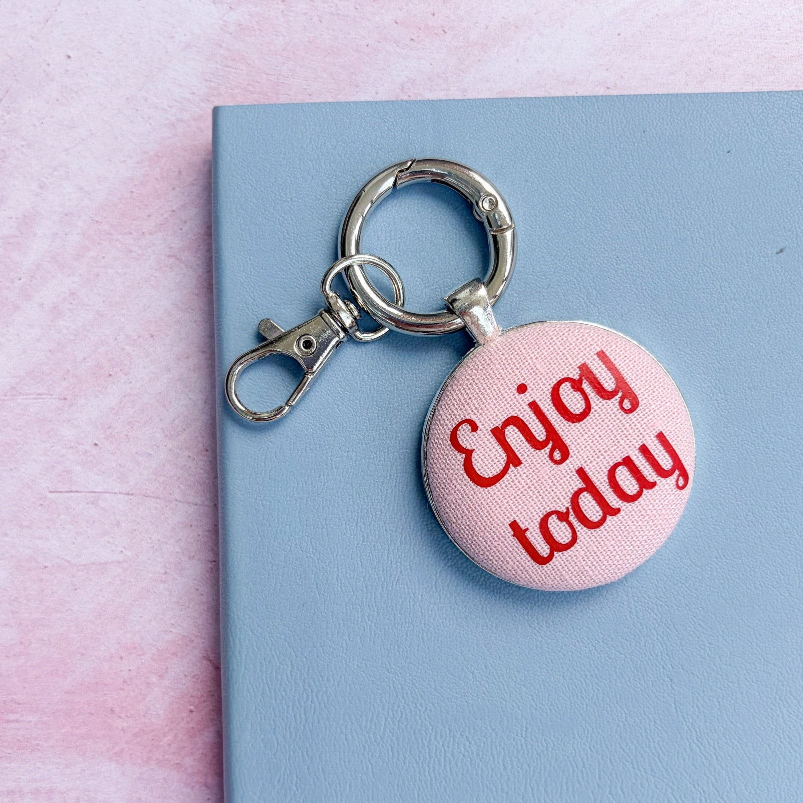 Pink keyring with 'Enjoy today' text printed in red on pink fabric, set on a blue surface
