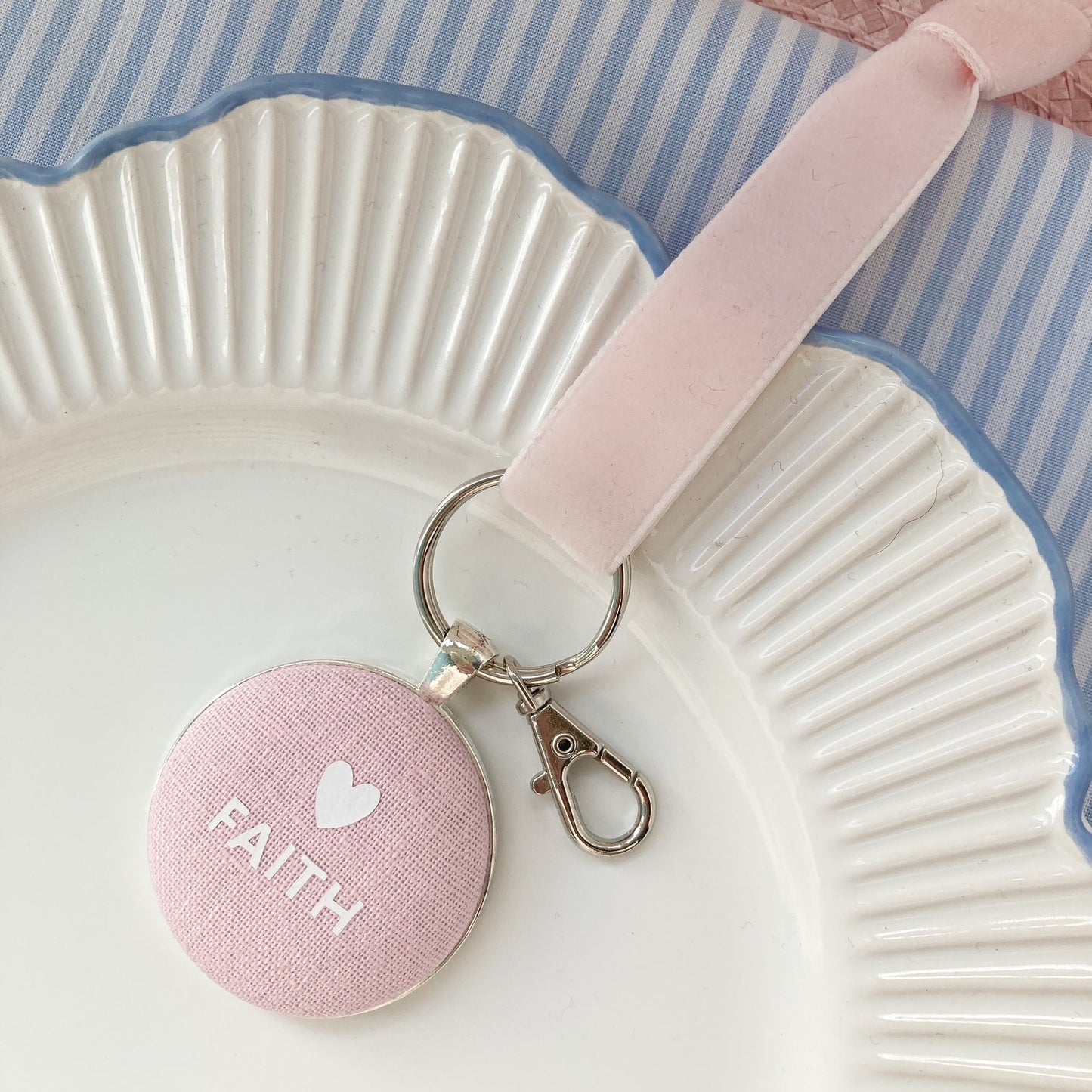 Pink fabric keepsake with printed 'FAITH' and a heart, on a white plate with blue rim