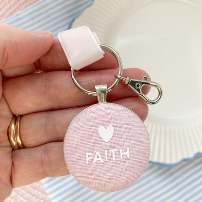 Pink fabric key ring with 'FAITH' and a heart symbol held in a hand against a striped background