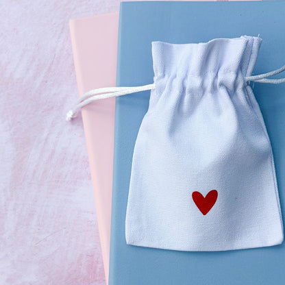 White drawstring bag with a red heart on a light blue box against a soft pink background