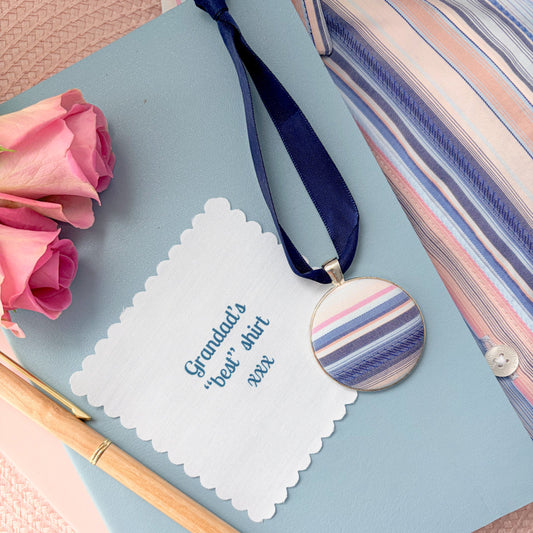 Memory fabric keepsake made with a striped shirt with a fabric message, placed on a blue notebook with a pen and pink flowers.