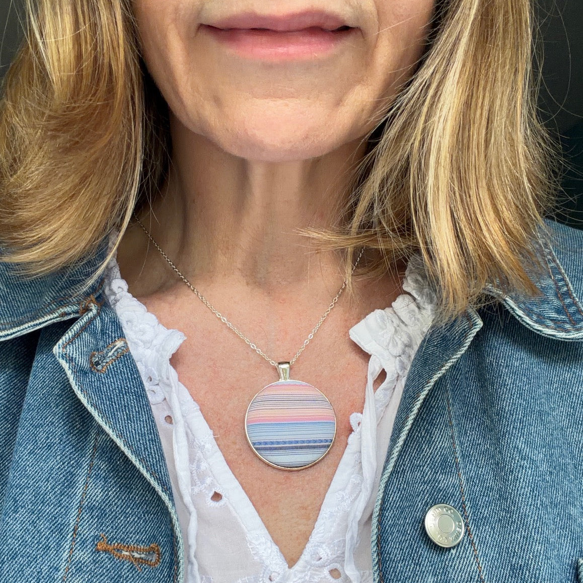 Person wearing a necklace with a circular striped pendant, dressed in a denim jacket and white blouse.