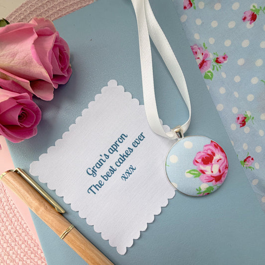 Grans apron memory keepsake thread on a ribbon, with a fabric message, set on a blue notebook with a pen and rose