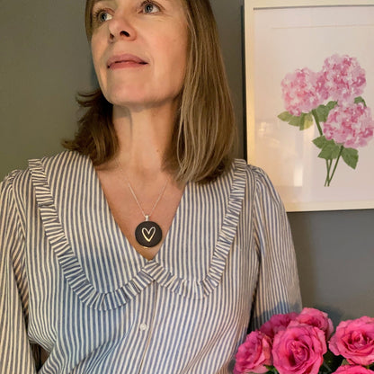 Person wearing a graphite grey necklace with heart design, with pink roses in a room with a floral painting on the wall.