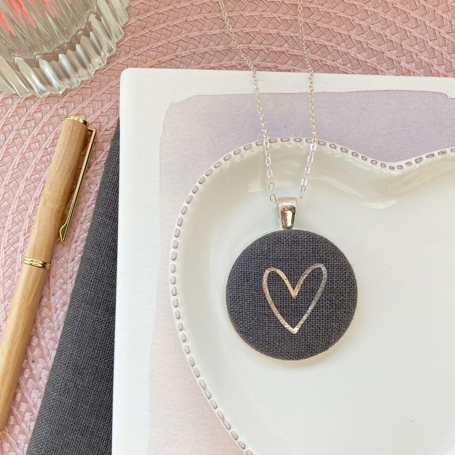 Heart-shaped pendant on a white dish with a pen and notebook on a pink surface