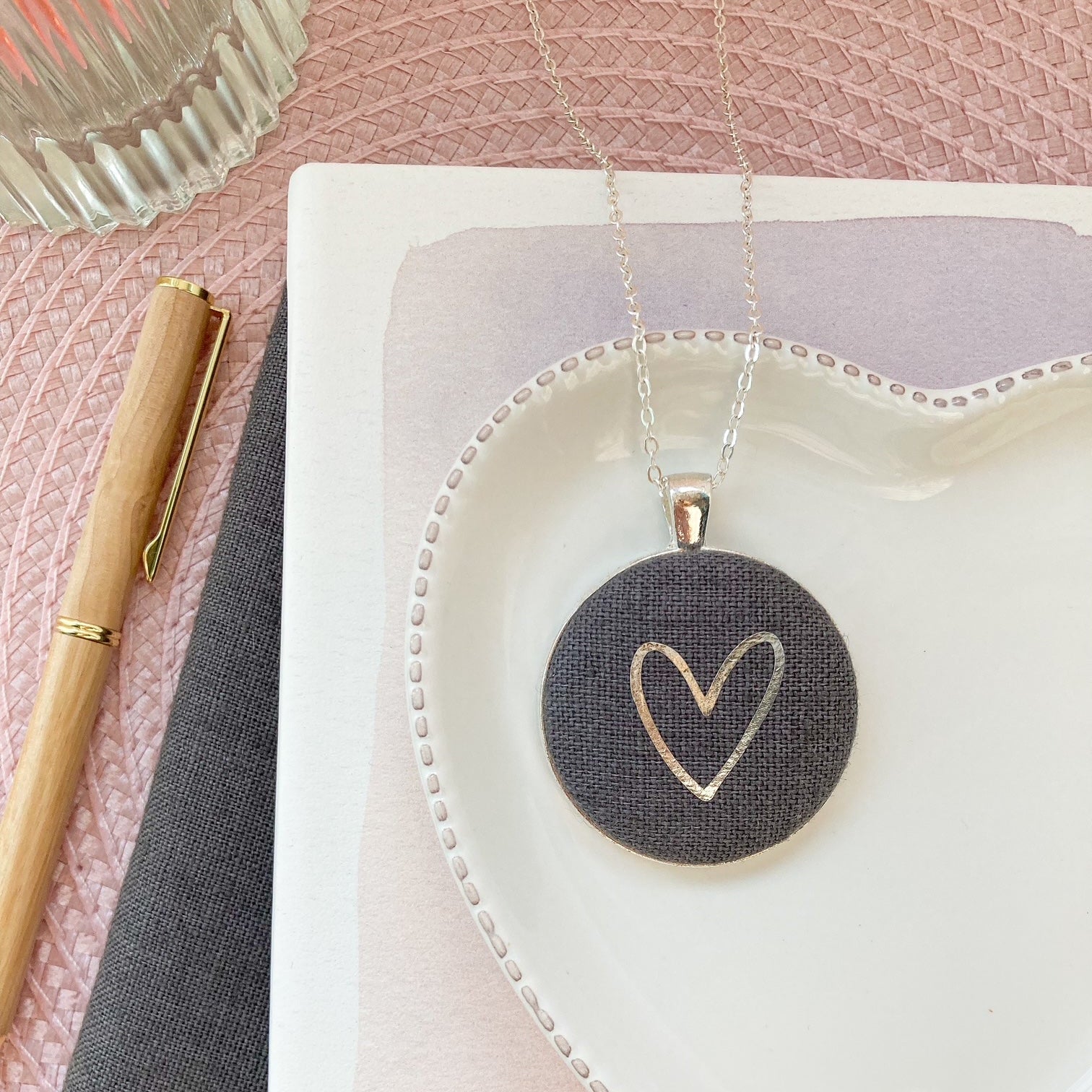 Heart-shaped pendant on a white dish with a pen and notebook on a pink surface