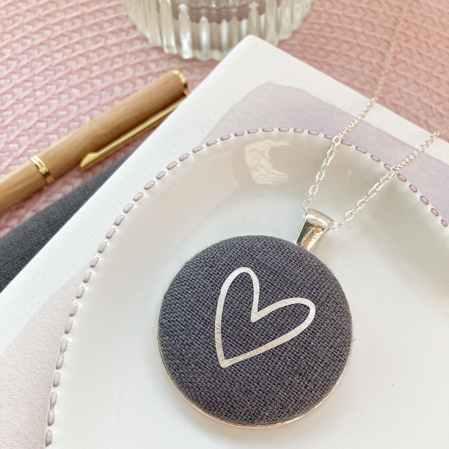 Dark grey necklace with heart pendant on a white dish with a pink background
