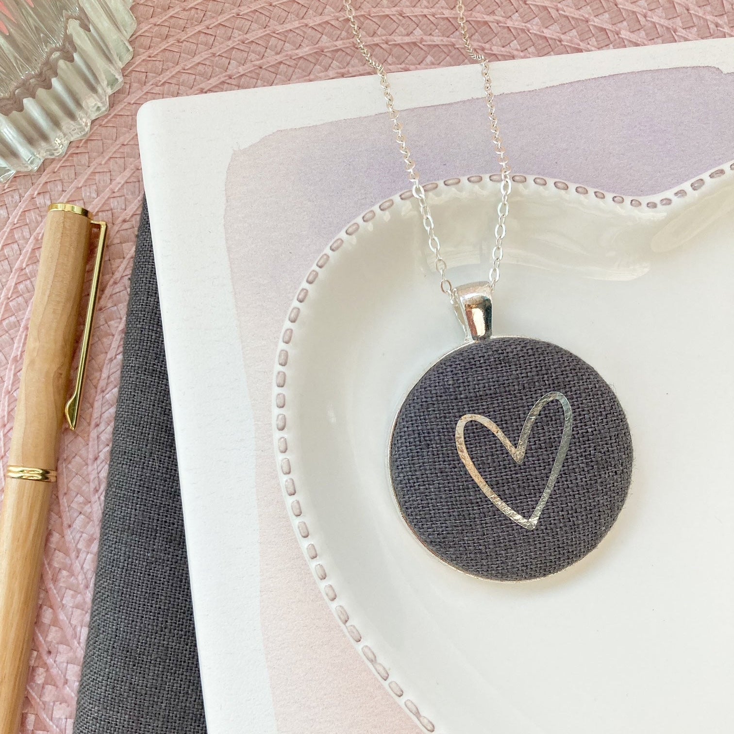 Dark grey necklace with a heart pendant on a white dish with a pink background