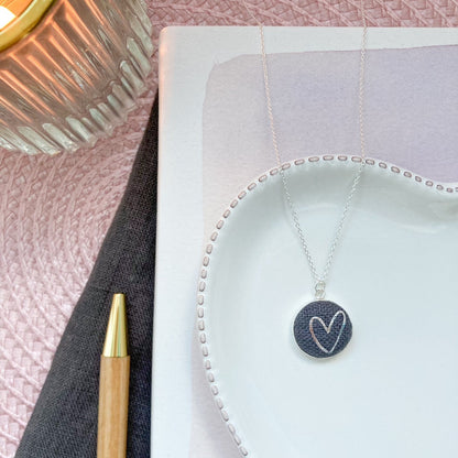 Jewelry necklace with heart pendant on a textured surface next to a pen and candle