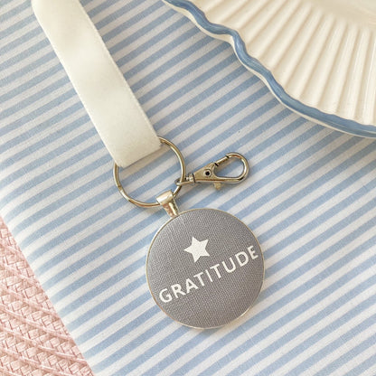Keychain with 'Gratitude' text and a star on grey fabric, on a striped fabric background
