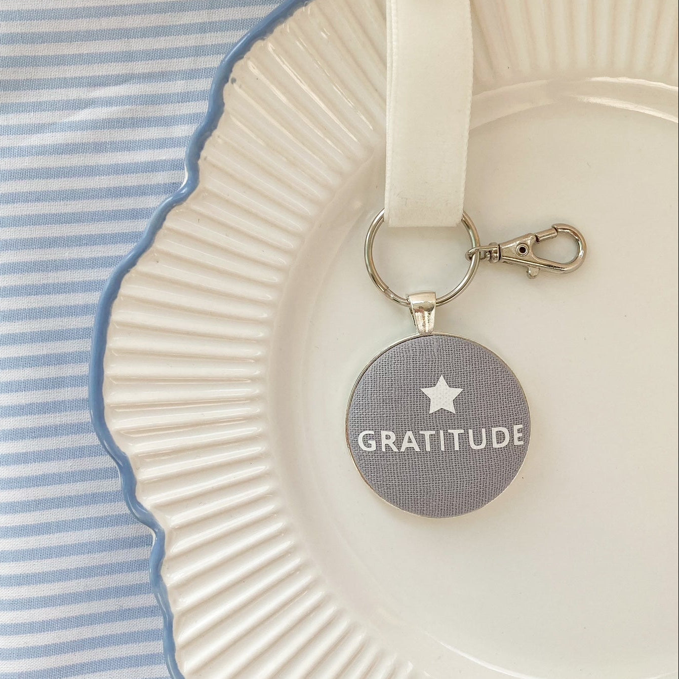 Keychain with 'Gratitude' laid on a white plate with blue rim, placed on a pink surface.
