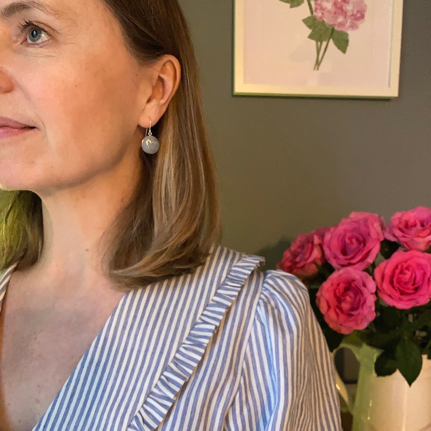 Person wearing a striped shirt and grey blue heart earrings with pink roses and a floral painting in the background
